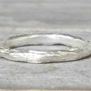 Può includere: Un anello d'argento con una superficie testurizzata dall'aspetto organico. L'anello è semplice e dal design moderno.