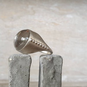 May include: A silver ring with a large, clear, rounded stone. The ring has a smooth, polished finish with a conical design and a row of small, decorative beads along the side. The ring is displayed on a gray, textured surface.