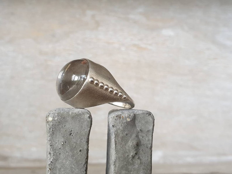 May include: A silver ring with a large, clear, rounded stone. The ring has a smooth, polished finish with a conical design and a row of small, decorative beads along the side. The ring is displayed on a gray, textured surface.