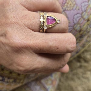 May include: Gold rings with gemstones. One ring features a triangular pink gemstone with a beaded border and two small diamonds. Another ring has a series of small gold circles. The rings are on a hand.