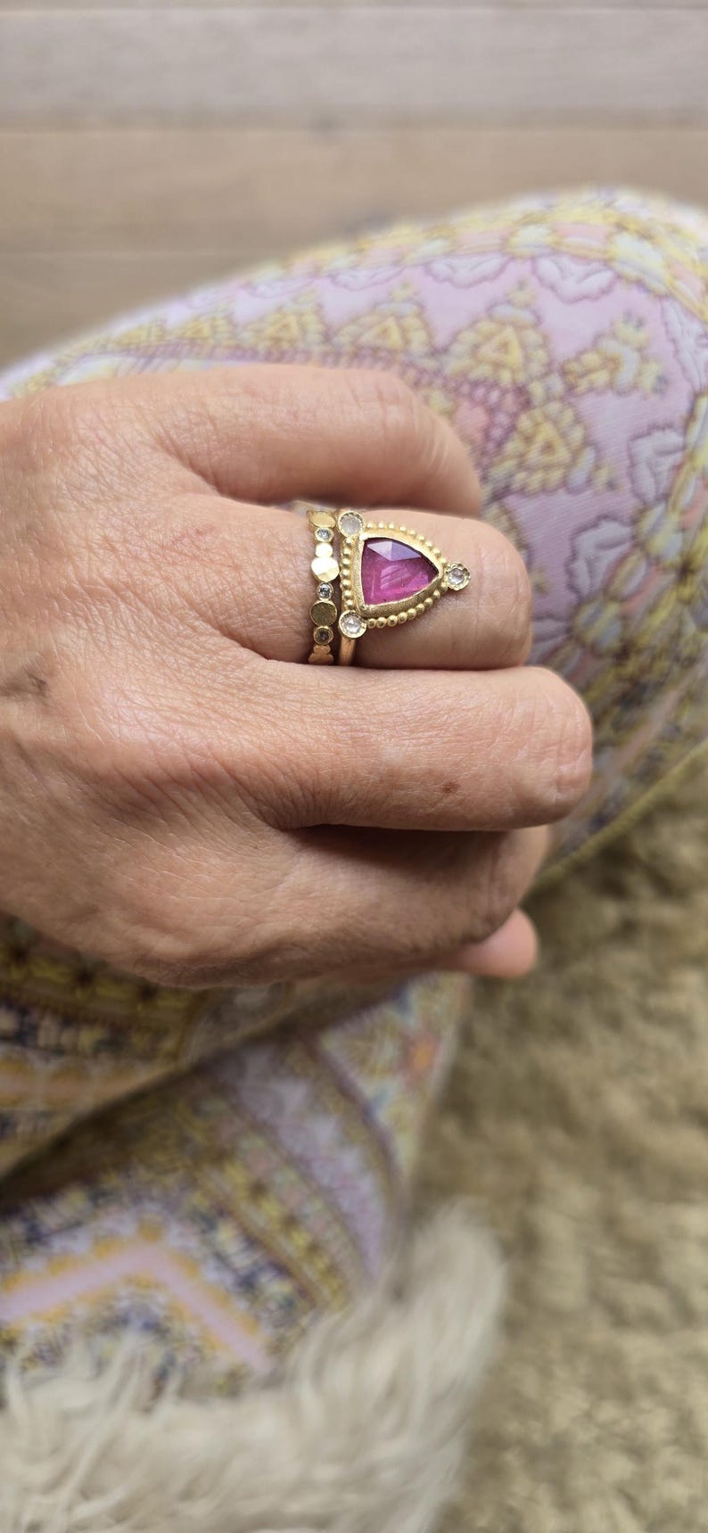 May include: Gold rings with gemstones. One ring features a triangular pink gemstone with a beaded border and two small diamonds. Another ring has a series of small gold circles. The rings are on a hand.