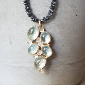 Pu&ograve; includere: Una delicata collana d'oro con un ciondolo che presenta un gruppo di pietre di acquamarina blu chiaro e piccoli diamanti trasparenti. La collana &egrave; realizzata con perline nere.