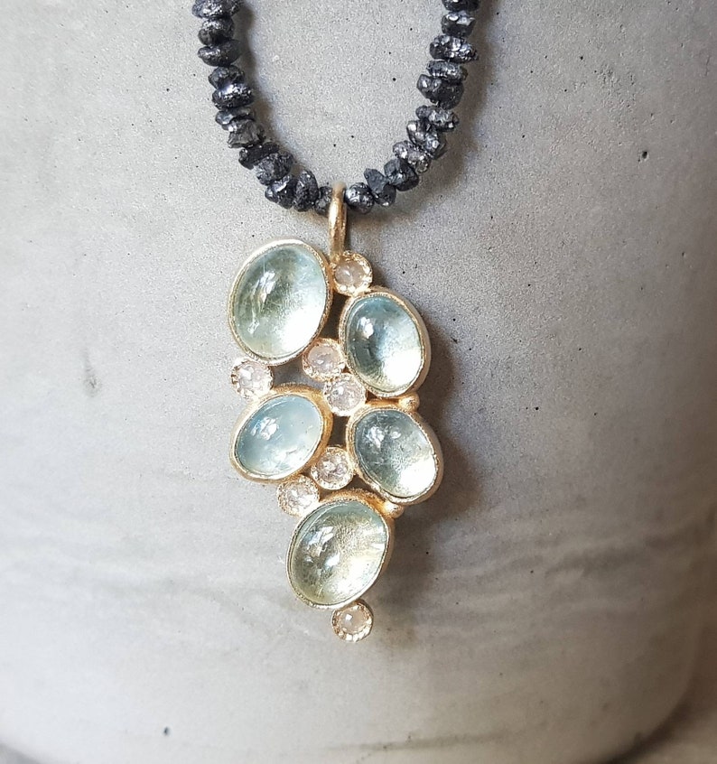 Pu&ograve; includere: Una delicata collana d'oro con un ciondolo che presenta un gruppo di pietre di acquamarina blu chiaro e piccoli diamanti trasparenti. La collana &egrave; realizzata con perline nere.