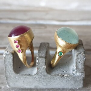 May include: Two gold rings with gemstone settings. The ring on the left has a deep red cabochon stone with three smaller pink stones on the side. The ring on the right has a light blue cabochon stone with a single green stone on the side.