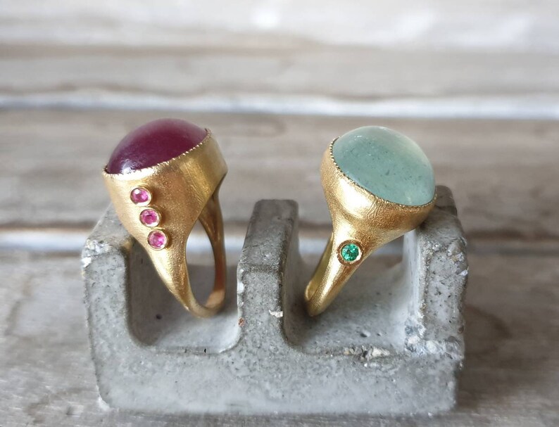 May include: Two gold rings with gemstone settings. The ring on the left has a deep red cabochon stone with three smaller pink stones on the side. The ring on the right has a light blue cabochon stone with a single green stone on the side.
