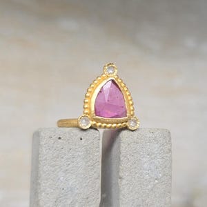 Puede incluir: Un anillo de oro con una gema rosa triangular, acentuada con pequeñas piedras blancas. El anillo tiene un diseño texturizado con cuentas alrededor de la piedra rosa. El anillo se muestra sobre una superficie gris texturizada.