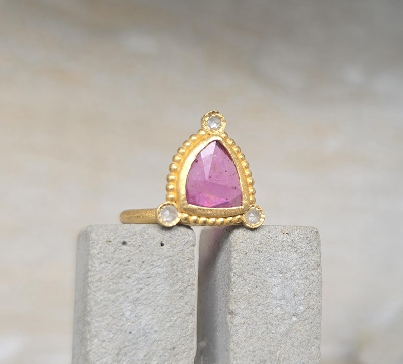 May include: A gold ring featuring a triangular pink gemstone, accented with small white stones. The ring has a textured, beaded design around the pink stone. The ring is displayed on a gray, textured surface.