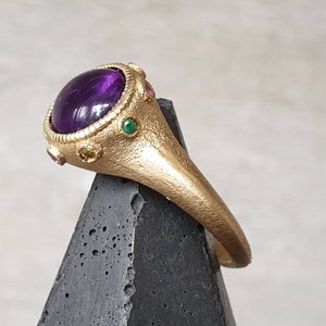 K&ouml;nnte beinhalten: Ein goldener Ring mit einem gro&szlig;en violetten Amethyst-Edelstein, umgeben von kleineren gr&uuml;nen und gelben Edelsteinen.