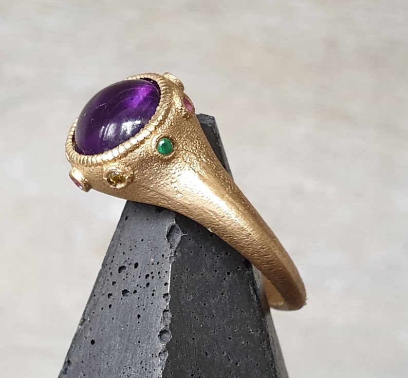 K&ouml;nnte beinhalten: Ein goldener Ring mit einem gro&szlig;en violetten Amethyst-Edelstein, umgeben von kleineren gr&uuml;nen und gelben Edelsteinen.
