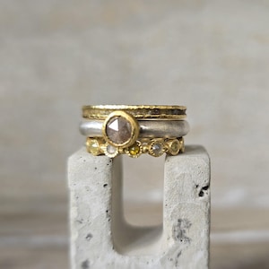 Pu&ograve; includere: Tre anelli d'oro con un diamante grigio sfaccettato pi&ugrave; grande nell'anello centrale. Gli anelli sono impilati su un blocco di cemento grigio.