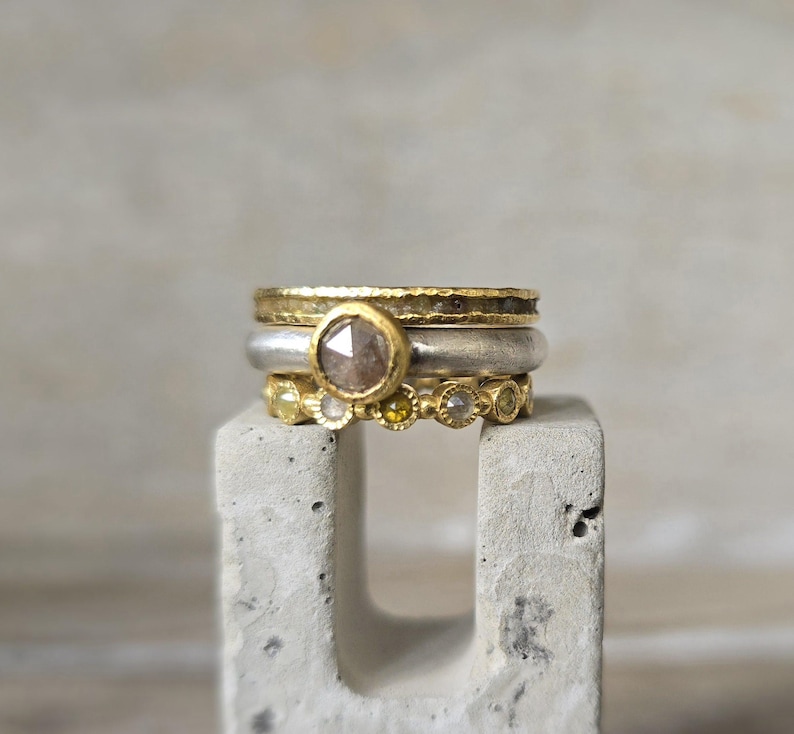 Pu&ograve; includere: Tre anelli d'oro con un diamante grigio sfaccettato pi&ugrave; grande nell'anello centrale. Gli anelli sono impilati su un blocco di cemento grigio.