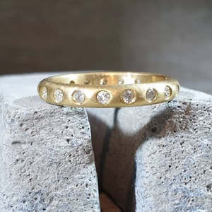 Peut inclure: Une bague dor&eacute;e avec une finition mate. La bague est orn&eacute;e d'une s&eacute;rie de petites pierres pr&eacute;cieuses claires serties autour du cercle. La bague est pr&eacute;sent&eacute;e sur une surface grise textur&eacute;e.