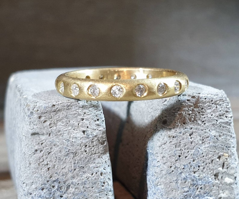 Peut inclure: Une bague dor&eacute;e avec une finition mate. La bague est orn&eacute;e d'une s&eacute;rie de petites pierres pr&eacute;cieuses claires serties autour du cercle. La bague est pr&eacute;sent&eacute;e sur une surface grise textur&eacute;e.