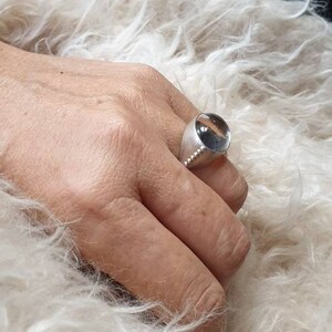May include: A silver ring with a large, clear, rounded stone. The ring has a wide band and a row of small, decorative beads. The ring is on a hand, with a fluffy, white background.