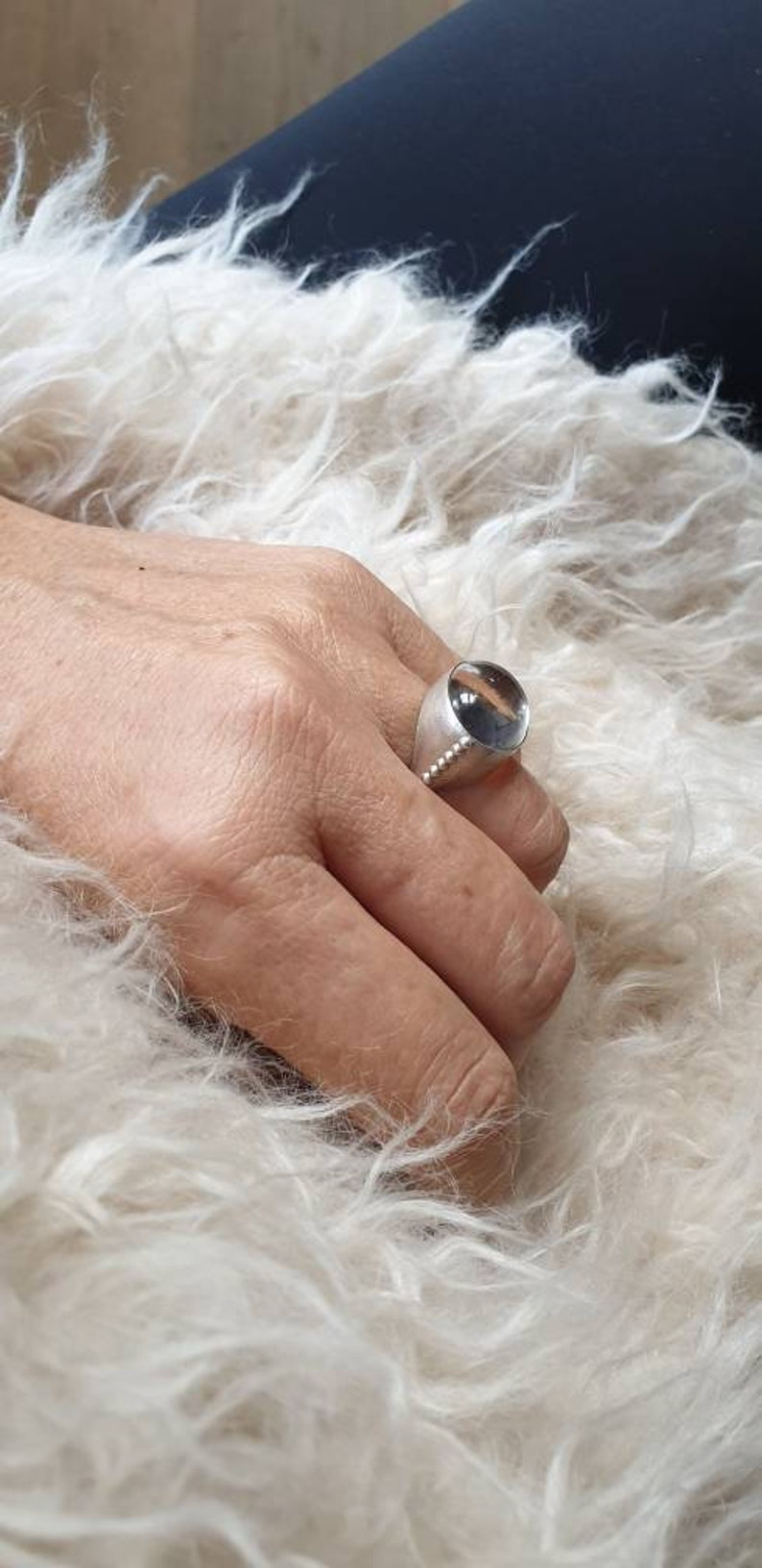 May include: A silver ring with a large, clear, rounded stone. The ring has a wide band and a row of small, decorative beads. The ring is on a hand, with a fluffy, white background.
