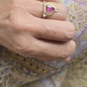 May include: A gold ring featuring a triangular, pink gemstone, flanked by two smaller, clear stones. The ring is set on a hand, with a patterned fabric visible in the background. The ring's design is simple yet elegant.