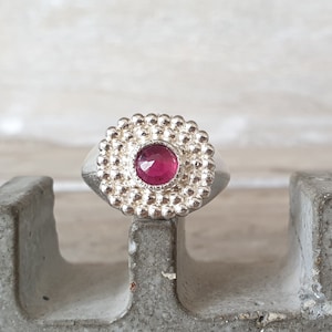 May include: A silver ring with a round, pink gemstone set in a beaded bezel. The ring is sitting on a gray concrete surface.