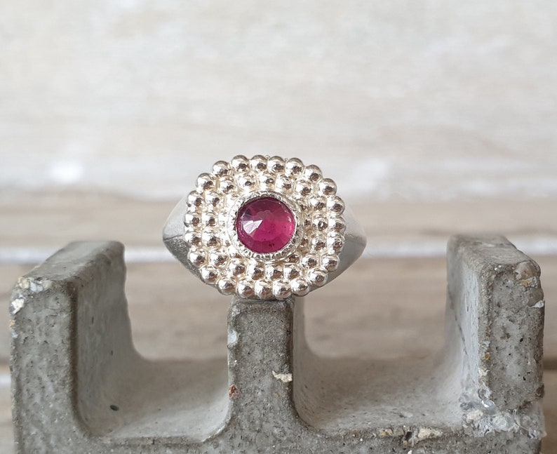 May include: A silver ring with a round, pink gemstone set in a beaded bezel. The ring is sitting on a gray concrete surface.