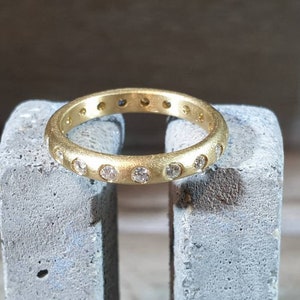 Peut inclure: Une bague en or avec une finition mate, sertie de nombreux petits pierres pr&eacute;cieuses transparentes. La bague est pr&eacute;sent&eacute;e sur une surface grise textur&eacute;e, mettant en valeur son design circulaire et l'&eacute;clat des pierres. La bande de la bague est d'une largeur constante.