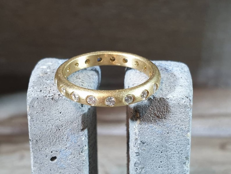 Peut inclure: Une bague en or avec une finition mate, sertie de nombreux petits pierres pr&eacute;cieuses transparentes. La bague est pr&eacute;sent&eacute;e sur une surface grise textur&eacute;e, mettant en valeur son design circulaire et l'&eacute;clat des pierres. La bande de la bague est d'une largeur constante.