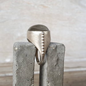 May include: A silver ring with a large, oval, smoky-colored stone. The ring has a textured band and a row of small, round embellishments along the side. The ring is displayed on a gray, textured surface.