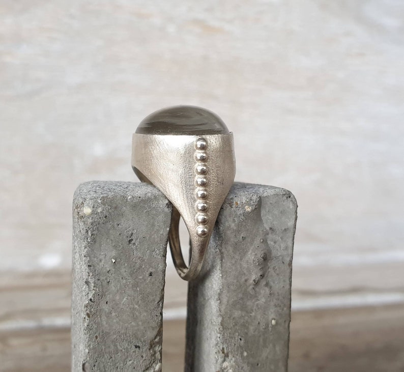 May include: A silver ring with a large, oval, smoky-colored stone. The ring has a textured band and a row of small, round embellishments along the side. The ring is displayed on a gray, textured surface.