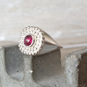 May include: A silver ring with a textured band and a single red gemstone in the center.