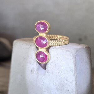 May include: A gold ring featuring three vertically aligned, oval-shaped, faceted pink rubies set in textured gold bezels. The ring band has a distinct beaded or granulated surface texture, providing a detailed and tactile appearance to the metalwork.