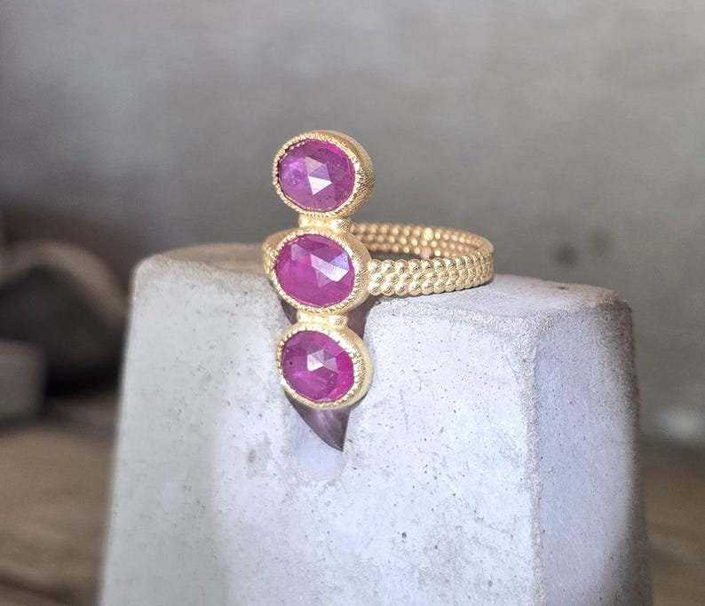 May include: A gold ring featuring three vertically aligned, oval-shaped, faceted pink rubies set in textured gold bezels. The ring band has a distinct beaded or granulated surface texture, providing a detailed and tactile appearance to the metalwork.
