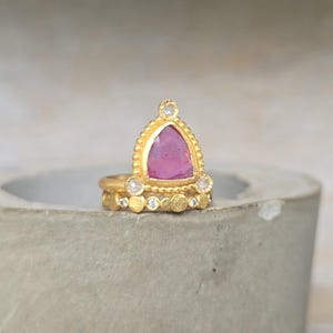 May include: A stack of gold rings with a pink gemstone and small diamonds. The top ring features a large, triangular pink stone set in a gold frame. The other rings have small diamonds and a textured design.