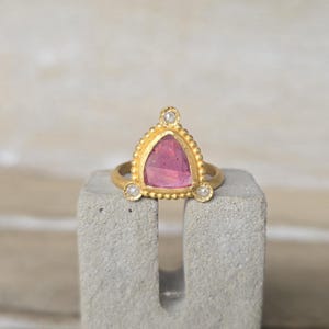 May include: A gold ring with a triangular pink gemstone and three small diamonds. The ring is set on a gray concrete block. The gemstone is a vibrant pink color, and the ring has a beaded border.