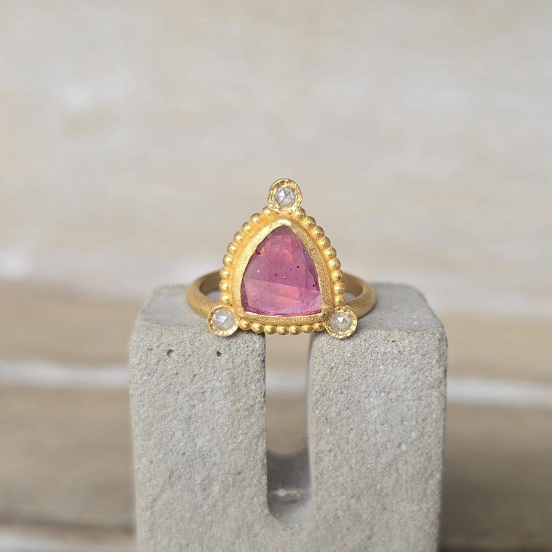 May include: A gold ring with a triangular pink gemstone and three small diamonds. The ring is set on a gray concrete block. The gemstone is a vibrant pink color, and the ring has a beaded border.