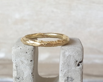 May include: A gold ring with a textured, organic design. The ring has a rough, bark-like appearance, with a band that is approximately 2 mm wide. The ring is displayed on a grey concrete block, showcasing its unique texture and colour.