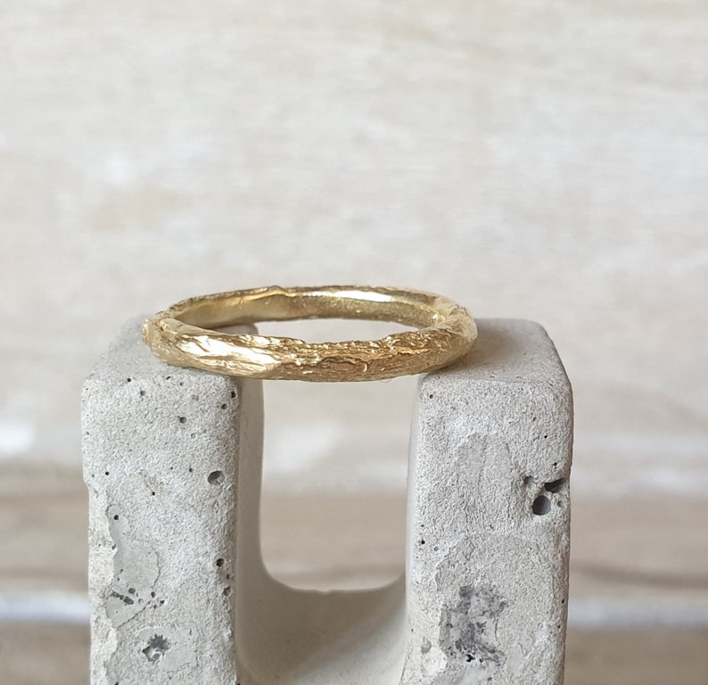May include: A gold ring with a textured, organic design. The ring has a rough, bark-like appearance, with a band that is approximately 2 mm wide. The ring is displayed on a gray concrete block, showcasing its unique texture and color.