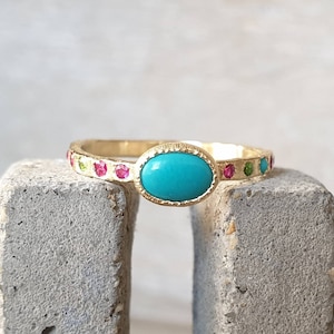 May include: A gold ring featuring a turquoise oval gemstone and a band of colourful gemstones. The ring is displayed on a grey concrete surface.