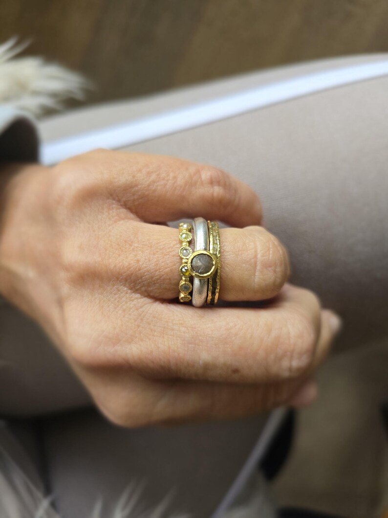 Pu&ograve; includere: Un set di tre anelli in oro e argento con un solo diamante rotondo nell'anello centrale. Gli anelli sono impilati su un dito.