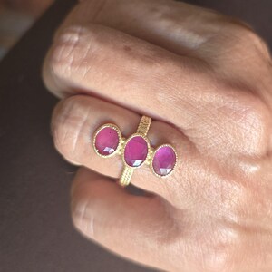 May include: A gold ring featuring three oval-shaped, faceted deep pink gemstones arranged horizontally. The band has a textured, cross-hatched pattern, and each gemstone is set in a gold bezel with a beaded edge detail surrounding the stones.