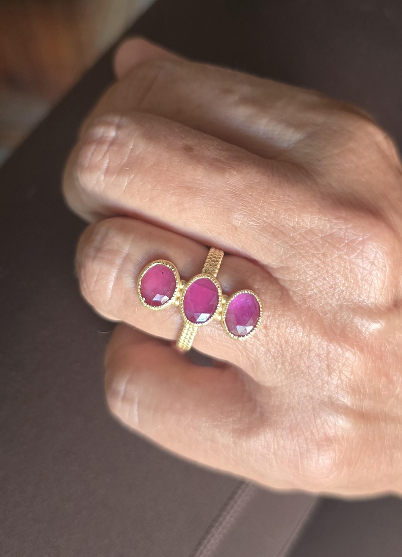 May include: A gold ring featuring three oval-shaped, faceted deep pink gemstones arranged horizontally. The band has a textured, cross-hatched pattern, and each gemstone is set in a gold bezel with a beaded edge detail surrounding the stones.