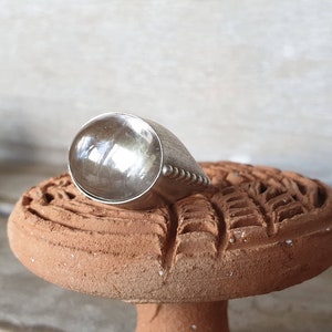 May include: A silver ring with a large, rounded top and a textured band. The ring is displayed on a terracotta-colored, carved surface. The ring's design is simple yet elegant, with a polished finish.