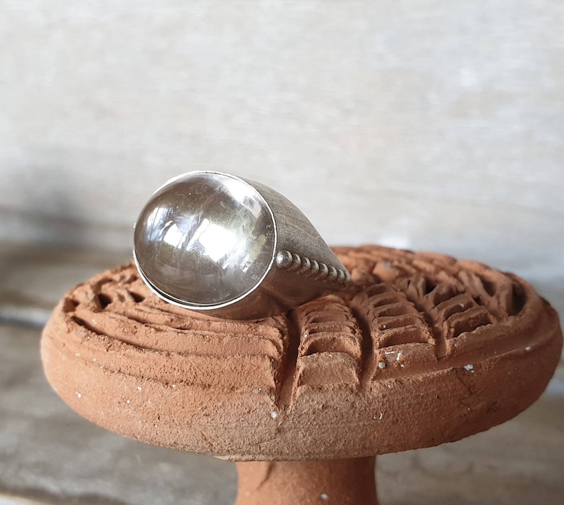 May include: A silver ring with a large, rounded top and a textured band. The ring is displayed on a terracotta-colored, carved surface. The ring's design is simple yet elegant, with a polished finish.