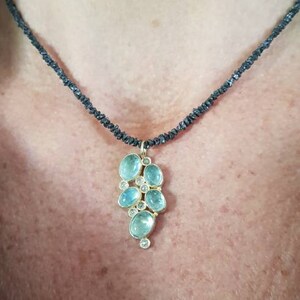 Pu&ograve; includere: Una collana delicata con un ciondolo che presenta un gruppo di pietre preziose di colore blu chiaro e trasparenti incastonate in oro. La collana &egrave; realizzata con perline nere.