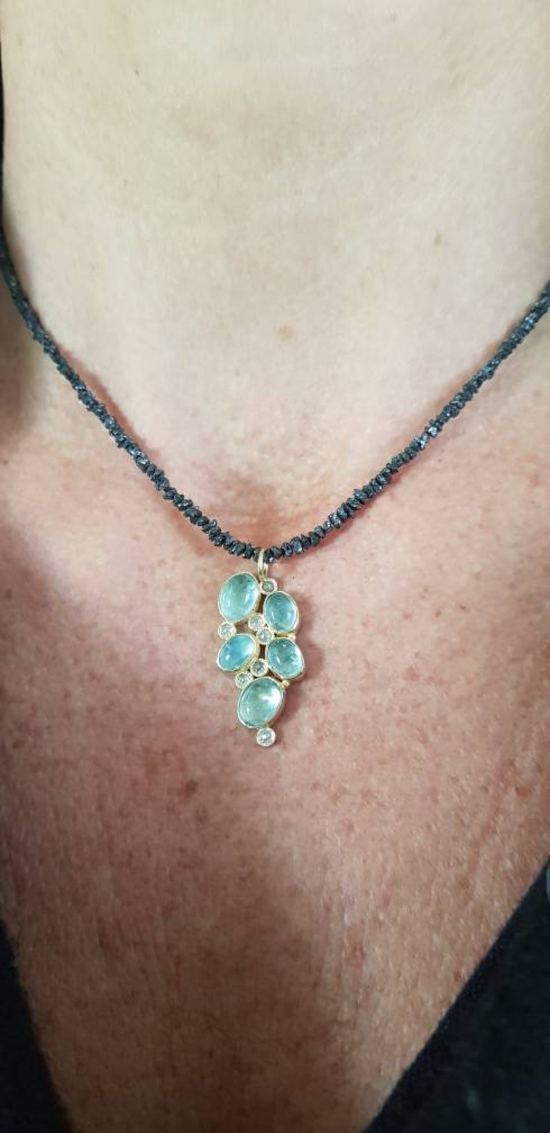 Pu&ograve; includere: Una collana delicata con un ciondolo che presenta un gruppo di pietre preziose di colore blu chiaro e trasparenti incastonate in oro. La collana &egrave; realizzata con perline nere.