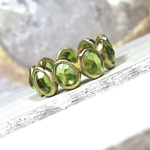 May include: A gold ring with a band of green peridot gemstones. The ring is set with oval-shaped stones that are bezel set in the gold.
