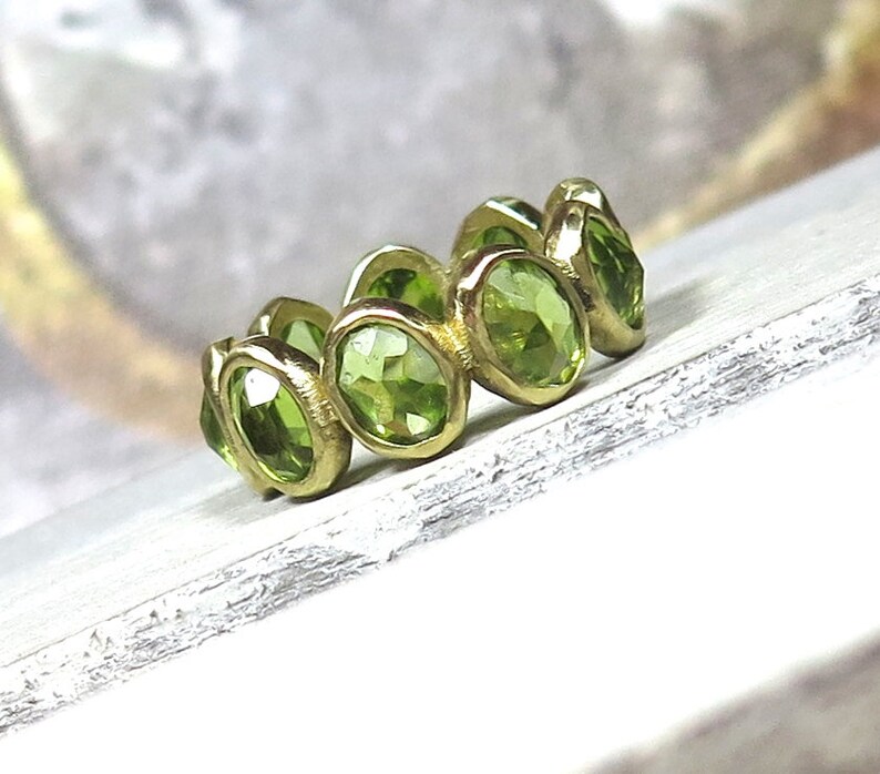 May include: A gold ring with a band of green peridot gemstones. The ring is set with oval-shaped stones that are bezel set in the gold.