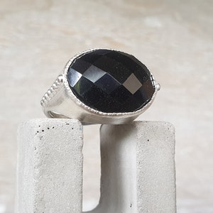 May include: A silver ring with a large, faceted black gemstone. The ring is set on a concrete block.