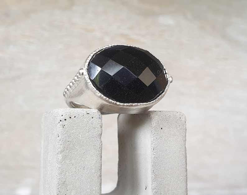 May include: A silver ring with a large, faceted black gemstone. The ring is set on a concrete block.