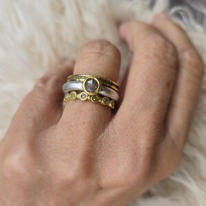 Pu&ograve; includere: Un set di tre anelli in oro e argento con una grande pietra preziosa grigia e pietre preziose verdi pi&ugrave; piccole. Gli anelli sono impilati su un dito.