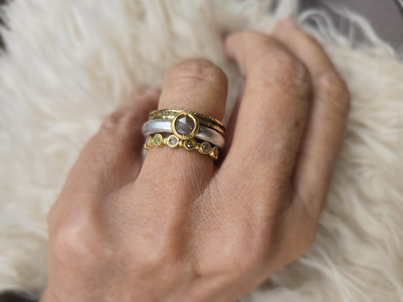 Pu&ograve; includere: Un set di tre anelli in oro e argento con una grande pietra preziosa grigia e pietre preziose verdi pi&ugrave; piccole. Gli anelli sono impilati su un dito.