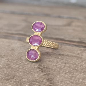 May include: A gold ring featuring three vertically aligned oval-shaped pink faceted gemstones. The band and the settings for the stones have a textured, granular surface finish. The stones are arranged in a straight line along the finger.