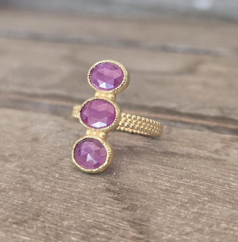 May include: A gold ring featuring three vertically aligned oval-shaped pink faceted gemstones. The band and the settings for the stones have a textured, granular surface finish. The stones are arranged in a straight line along the finger.